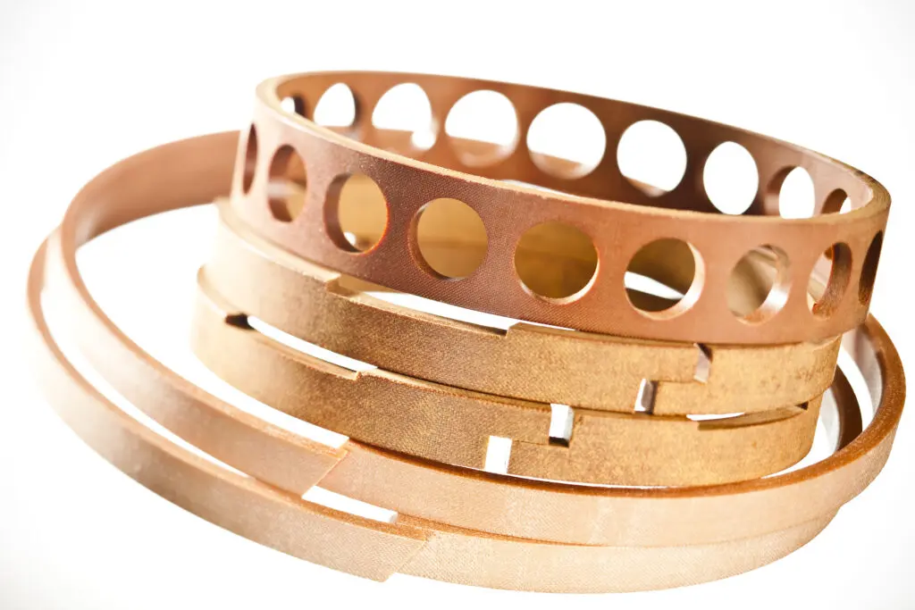 Close-up of several brown, circular metal bands with holes and slots, stacked together on a white background, shown alongside Cotton Fabric Laminates SRBF for comparison.