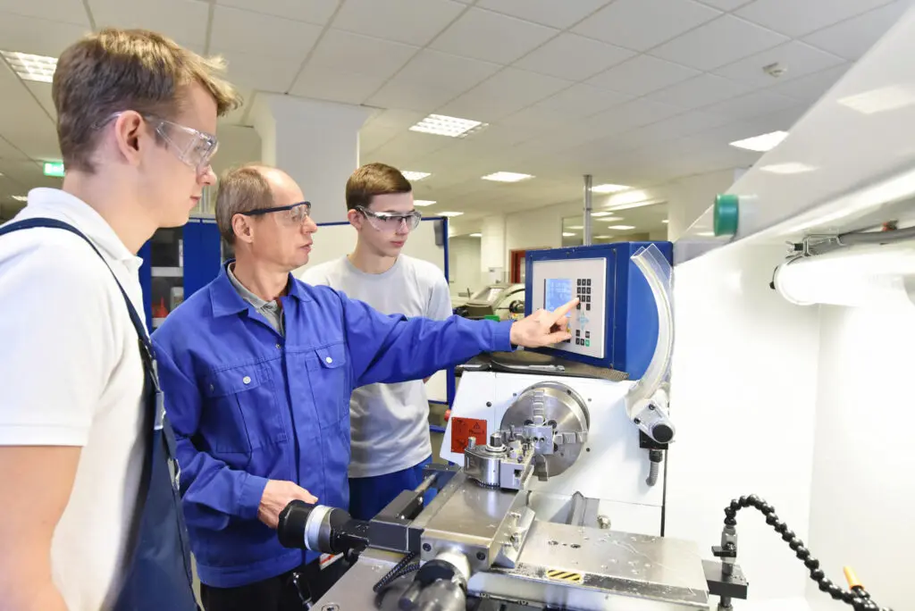 Three men in safety goggles operate and discuss cnc machining at a CNC machine in a bright, modern workshop.