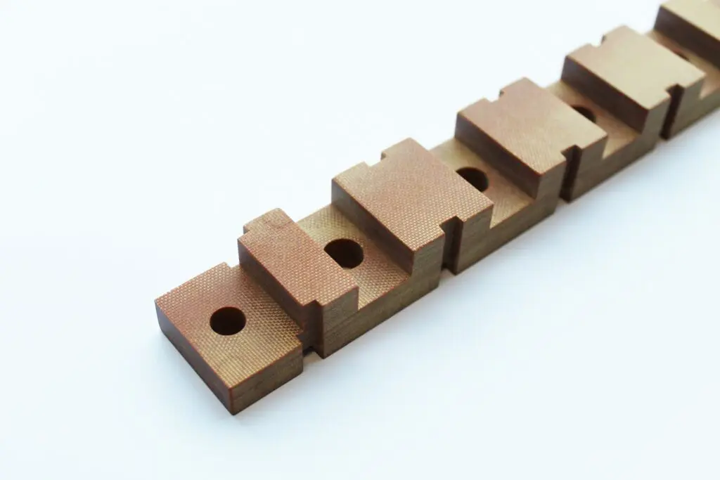 A row of rectangular, copper-colored SRBGF Material metal blocks with holes and notches, arranged diagonally on a white background.