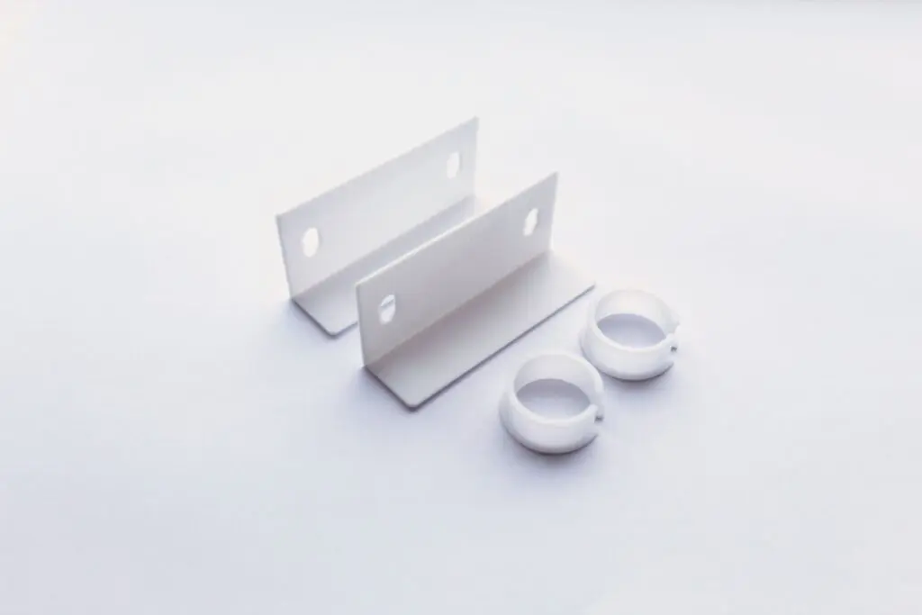 Two white metal brackets with holes and two white plastic circular clips, made from thermoplastic materials, are displayed on a white surface.