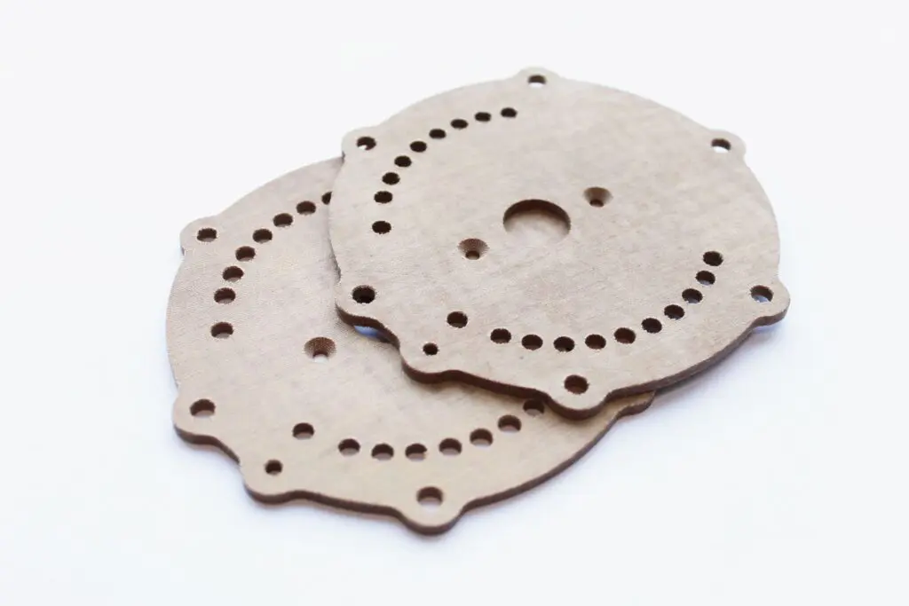 Two round, flat metal plates with holes and notches, resembling components used in fabric based laminates, are shown overlapping on a white background.