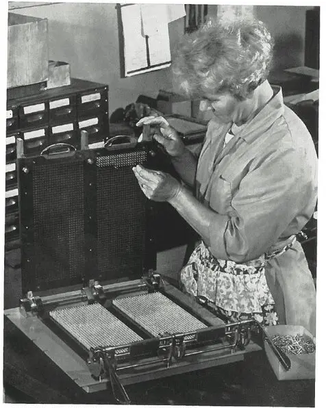 A woman inserts metal pins into a large grid-like device in an office or laboratory setting.