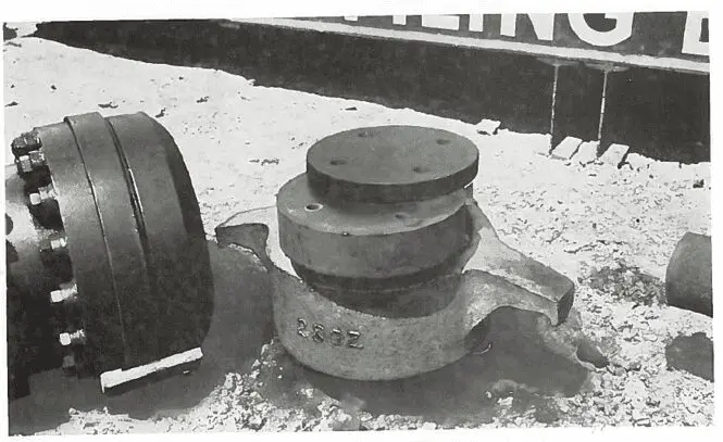 A large metal valve assembly sits on sandy ground near industrial equipment.