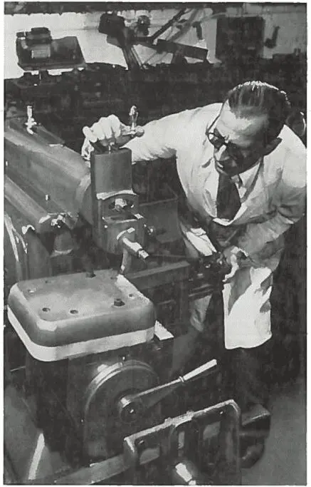 Man in a lab coat operates or inspects a large industrial machine in a workshop or laboratory.