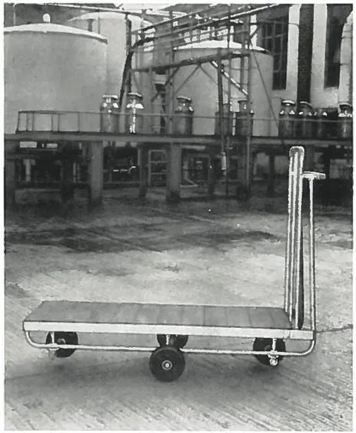 A flatbed cart with three wheels stands on concrete in an industrial facility with large tanks in the background.