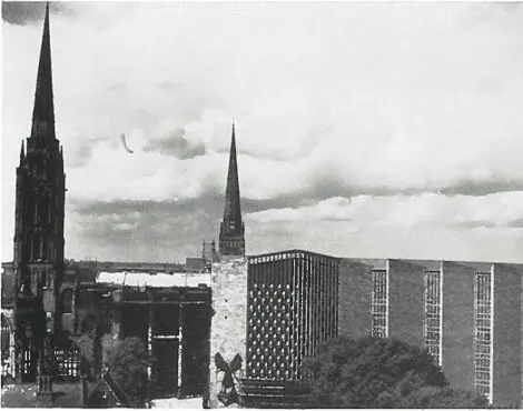 Two church spires and a modern building rise above trees and rooftops under a cloudy sky.