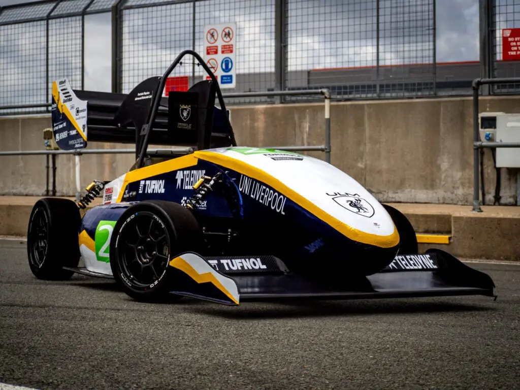 Blue and white race car with UNI LIVERPOOL logo parked on a racetrack near a concrete barrier.