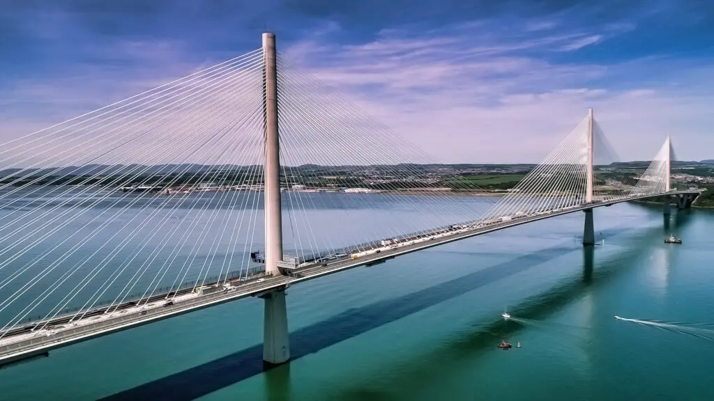 A modern cable-stayed bridge spans a wide river under a bright blue sky with scattered clouds.
