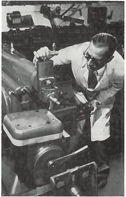 Man in a lab coat operates or inspects a large industrial machine in a workshop or laboratory.