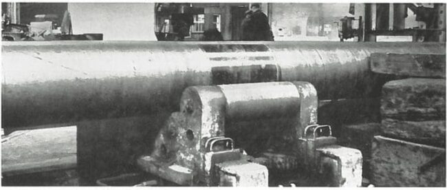 Large metal cylinder supported by heavy machinery in an industrial workshop, with workers in the background.