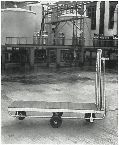 A flatbed cart with three wheels stands on concrete in an industrial facility with large tanks in the background.