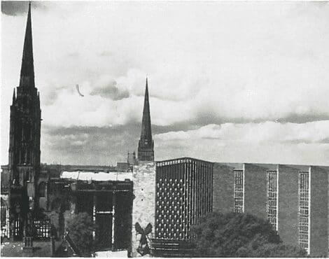 Two church spires and a modern building rise above trees and rooftops under a cloudy sky.