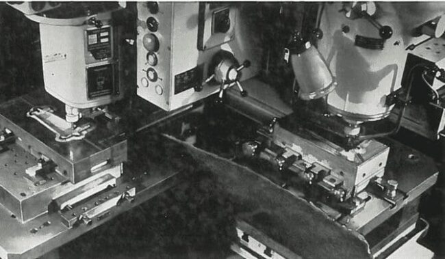 Two industrial machines with control panels and clamps, processing metal parts in a manufacturing setting.