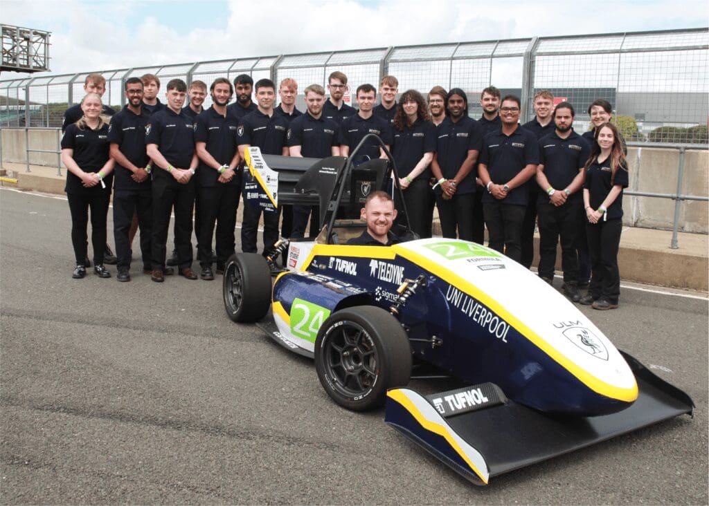 A team of students poses behind a race car on a track, with one person seated in the car.